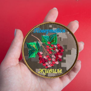May include: Round embroidered patch with a camouflage background and a floral design featuring red berries and green leaves. The patch has blue and yellow text. The patch is held in a hand against a red background.