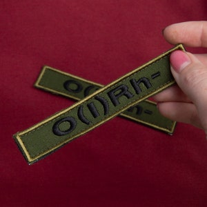 May include: Two olive green rectangular patches with black embroidered text. The text reads "O(I)Rh-". One patch is held by a hand with pink nail polish. The patches have a thin gold border and are set against a deep red background.
