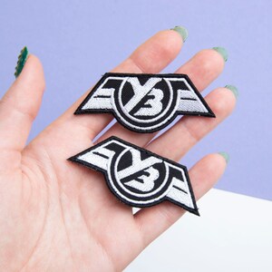 Puede incluir: Dos parches bordados en blanco y negro. Cada parche presenta un emblema estilizado con las letras "YB" dentro de un círculo, flanqueado por formas de ala. Los parches se sostienen en una mano sobre un fondo morado y blanco.