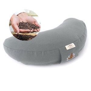 May include: A gray crescent-shaped yoga bolster with a stitched seam. A small inset shows hands holding buckwheat hulls. The bolster has a tag with a tree logo and the text "NATURBUDA".