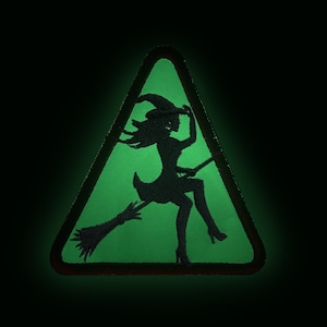May include: A black patch with a green triangle background featuring a silhouette of a witch riding a broom. The witch is wearing a hat and dress. The patch has a black border and is set against a dark background.