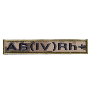 May include: A rectangular patch with the text "AB(IV)Rh+" embroidered in dark gray thread. The background has a camouflage pattern in shades of brown and tan. The patch is bordered with a thin, olive green stitching.
