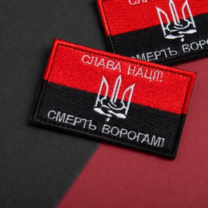 May include: Embroidered rectangular patch in red and black. The top section is red, the bottom black, with a white trident emblem. The text "Glory to the Nation!" and "Death to Enemies!" are embroidered in white.