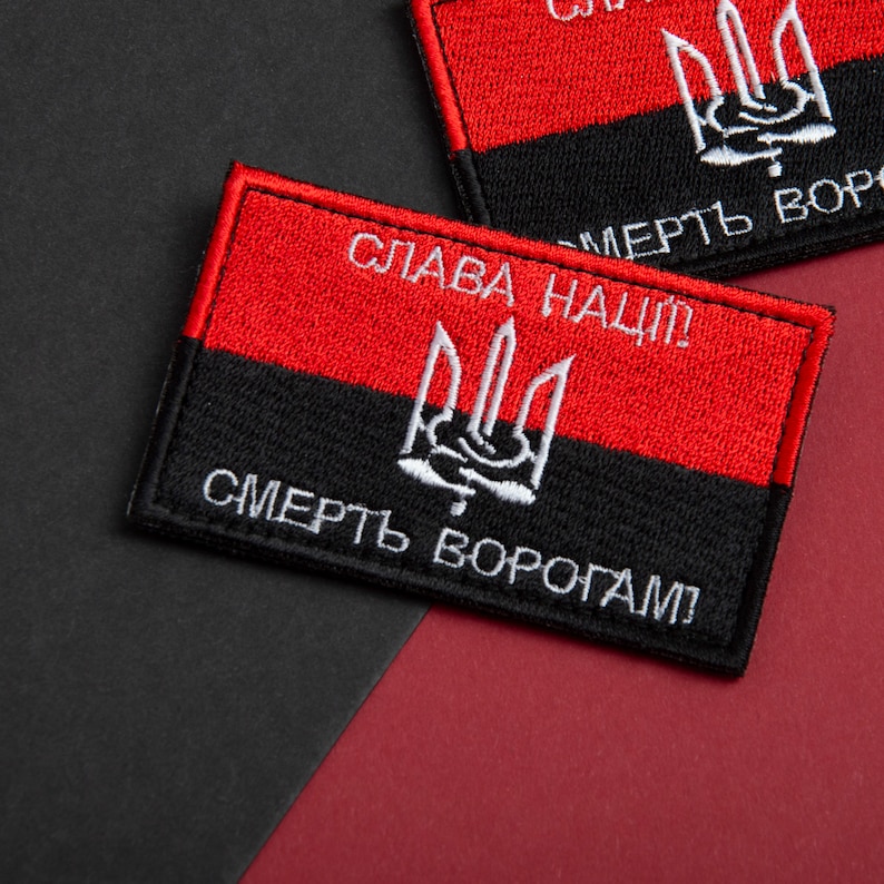 May include: Embroidered rectangular patch in red and black. The top section is red, the bottom black, with a white trident emblem. The text "Glory to the Nation!" and "Death to Enemies!" are embroidered in white.