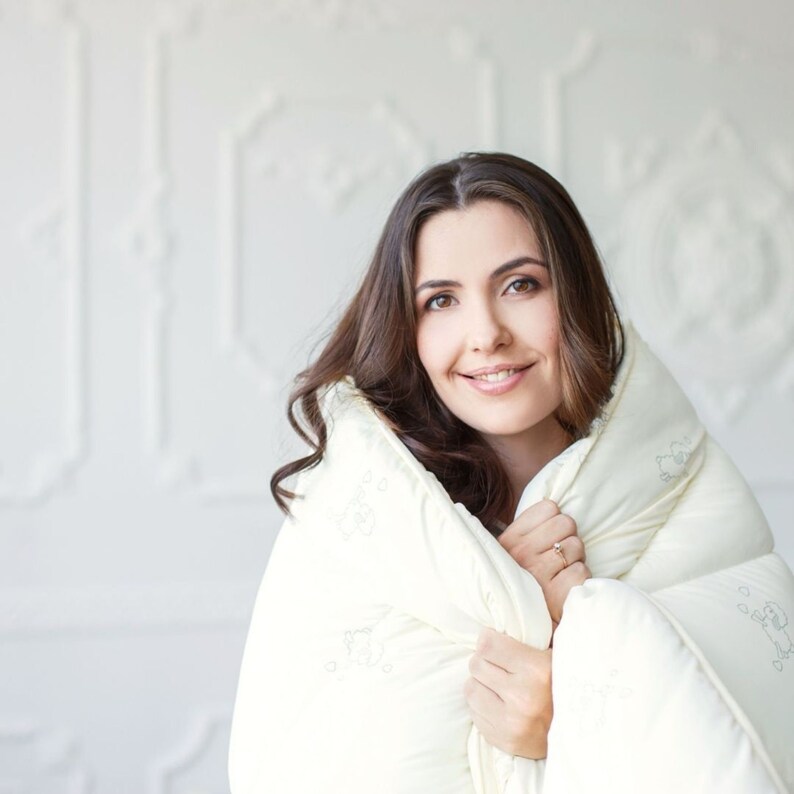 Puede incluir: Una mujer envuelta en una manta acolchada de color crema. La manta tiene un patr&oacute;n sutil y parece suave y c&oacute;moda. La mujer sonr&iacute;e, con el pelo largo y casta&ntilde;o enmarcando su rostro. El fondo es una pared blanca con molduras decorativas.