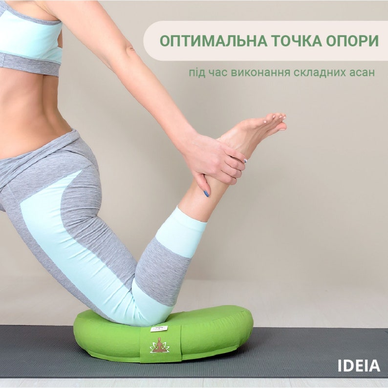 May include: A green yoga bolster with a light green strap and a logo. The bolster is on a dark gray mat. The text on the image says "Optimal support point" and "during complex asanas".