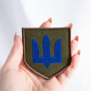 May include: A square patch featuring the Ukrainian coat of arms, a blue trident, embroidered on a green background. The patch has a brown border and is held in a hand against a white backdrop.