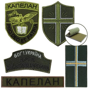 May include: A collection of olive green embroidered patches. One patch features a shield shape with a cross design. Another patch has the word "KAPEAHAH" in a banner shape. Other patches have text and a bird design.