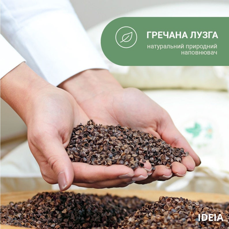 May include: Close-up of hands holding a pile of buckwheat hulls. The hulls are dark brown and appear to be a natural product. The image includes text in Ukrainian that translates to "buckwheat husk, natural natural filler."