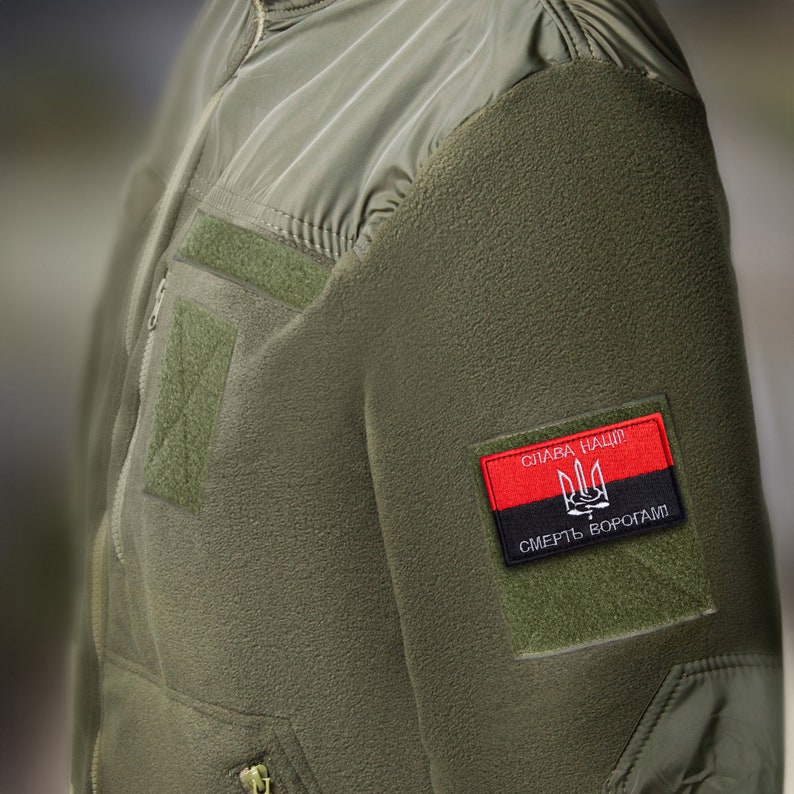 May include: An olive green jacket with a black and red patch. The patch displays a trident symbol and the text "Glory to the Nation! Death to enemies!" The jacket has a zip front and several velcro patches.