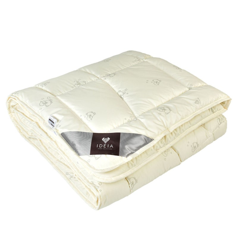 Puede incluir: Manta acolchada de color crema con un patr&oacute;n repetido de peque&ntilde;as formas grises en forma de nube. La manta est&aacute; doblada, mostrando su textura y dise&ntilde;o. Una etiqueta con el texto "IDEIA HOME COLLECTION" es visible.