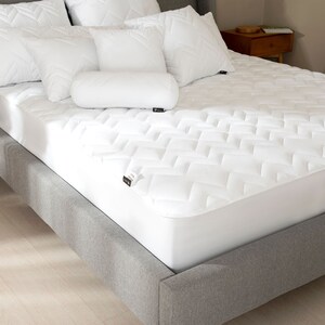 Peut inclure: Un lit avec un protège-matelas et des oreillers blancs. Le protège-matelas présente un motif géométrique matelassé. Plusieurs oreillers blancs de différentes formes, dont carrés et cylindriques, sont disposés sur le lit. Le cadre de lit est gris.