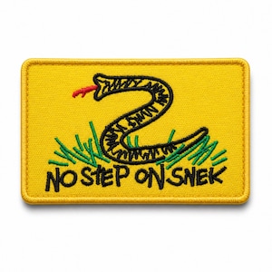 May include: A rectangular yellow embroidered patch featuring a black snake with a red tongue, green grass blades, and the black text NO STEP ON SNEK. The patch has a yellow stitched border and a flat, textured surface design.