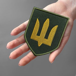 May include: A green and gold embroidered patch featuring a trident design. The patch has a dark green background with a gold trident emblem in the center. The patch is shield-shaped with a green border.