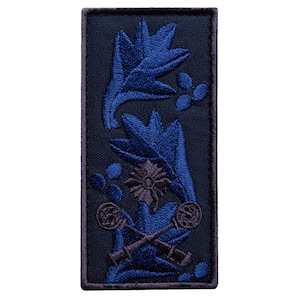 Pode incluir: Um patch retangular com fundo azul marinho escuro. O patch apresenta desenhos florais azuis bordados, incluindo folhas e um emblema central em forma de estrela. Dois canhões cruzados na parte inferior.