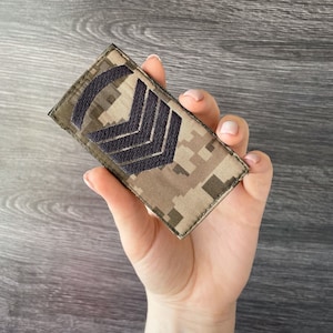 May include: A rectangular patch with a camouflage pattern in shades of brown, tan, and green. Three black chevrons are embroidered on the patch. The patch is held in a hand.