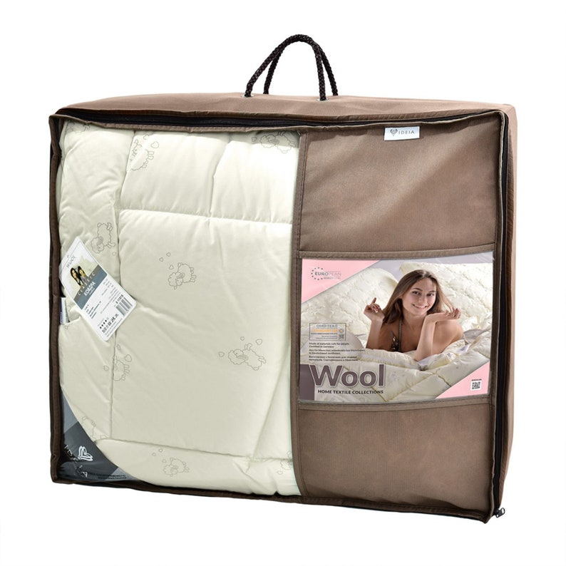 Puede incluir: Un juego de edred&oacute;n beige en una bolsa de transporte marr&oacute;n. El edred&oacute;n es de color crema con un patr&oacute;n sutil. La bolsa de transporte tiene un asa y una ventana transparente que muestra el edred&oacute;n y una mujer. La palabra "Wool" es visible.