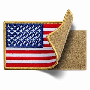 May include: A rectangular embroidered patch featuring the United States flag with red, white, and blue threads. The patch has a gold-colored border and is partially peeled back to reveal a tan-colored hook-and-loop fastener backing.
