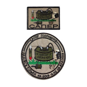 May include: Two embroidered patches. The top patch is rectangular with the word "SAPER" and a green and brown design. The bottom patch is circular with text and a similar design. Both patches feature a military theme.