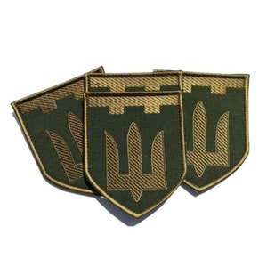 May include: A collection of olive green shield-shaped patches with a gold embroidered trident design. The patches have a gold border and a textured appearance, likely for application on clothing or gear.