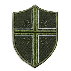 May include: A shield-shaped patch with a cross design. The patch is primarily dark green with a lighter green cross and white accents. The cross design has radiating lines in each quadrant.
