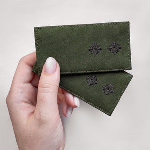 Puede incluir: Dos parches de tela verde oliva con diseños florales bordados en marrón oscuro. Uno es rectangular y el otro triangular. Los parches son sostenidos por una mano sobre un fondo blanco.