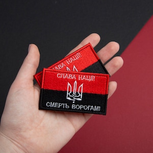 May include: Two rectangular embroidered patches in red and black. The top half is red with white text, the bottom half black with white text. A white trident emblem is centered on the black section. The patches are held in a hand.