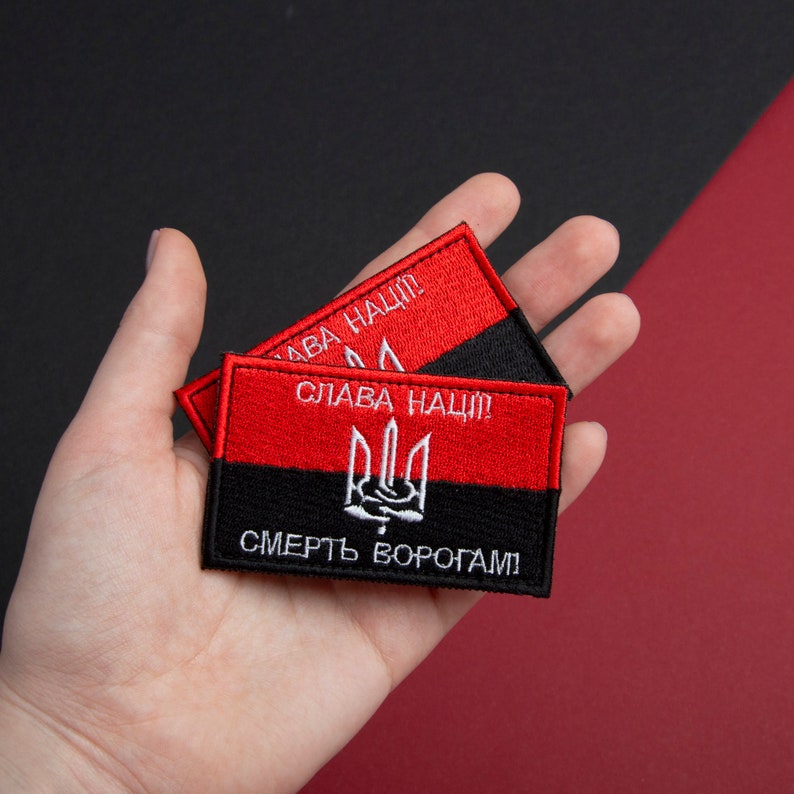 May include: Two rectangular embroidered patches in red and black. The top half is red with white text, the bottom half black with white text. A white trident emblem is centered on the black section. The patches are held in a hand.