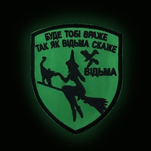 May include: A green patch with black text and a witch silhouette riding a broom. The patch features the text "BUДE TOБI BPAЖE TAK ЯK BIДЬMA CKAЖE" and the word "BIДЬMA". A black cat and two birds are also depicted.