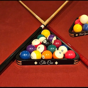 May include: A black wooden pool ball rack with the text "The Cue" on the front. The rack is filled with 15 billiard balls, including the cue ball, in a triangular formation. The rack is on a green felt pool table with a brown wooden cue stick crossing over the top.