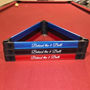May include: A stack of three pool ball racks in blue, black, and red. Each rack is triangular and has the text "Behind the 8 Ball" in white script. The racks are stacked on a red felt pool table.