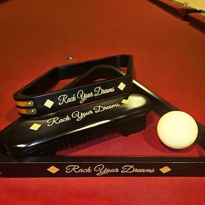 May include: A black wooden billiard ball rack and a matching black cleaning brush, both featuring gold diamond accents and the white text "Rack Your Dreams". A single white billiard ball is positioned inside the triangular rack.