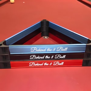 May include: A stack of three triangular billiard ball racks in blue, black, and red. Each rack features the white text "Behind the 8 Ball" in a cursive script font on the front side. The racks are constructed with black corner joints.