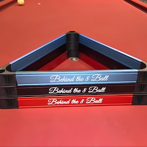 May include: A stack of three triangular billiard ball racks in light blue, black, and red. Each rack features the white script text Behind the 8 Ball printed on the front side. The racks are constructed with black corner joints and colored rails.