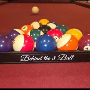 May include: A black plastic billiard ball rack containing a full set of colorful numbered pool balls arranged in a triangle. The front of the rack features the white text Behind the 8 Ball. A single white cue ball sits on the red felt table surface.