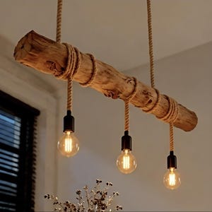 Può includere: Una lampada a sospensione composta da un tronco di legno naturale orizzontale sospeso da una spessa corda color marrone chiaro. Tre portalampade neri sono avvolti attorno al tronco con corda, ciascuno con una lampadina rotonda trasparente.