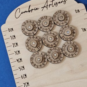 Könnte beinhalten: Ein hölzernes Messwerkzeug mit kreisförmigen Holzstücken, die mit Zahlen markiert sind. Das Werkzeug hat sowohl Zoll- als auch Zentimeter-Markierungen. Der Text "Cumbria Artisans" steht oben.