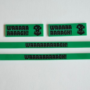 Op de afbeelding: Groene rechthoekige stukken met de tekst "WAAAAAAAGH!" in zwart. Elk stuk heeft een schedelafbeelding. De stukken variëren in grootte en zijn gerangschikt op een wit oppervlak.