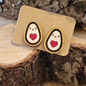 Può includere: Orecchini a bottone in legno a forma di avocado. Ogni orecchino presenta un volto sorridente e un cuore rosso. Gli orecchini sono su un cartoncino marrone, su uno sfondo di legno.