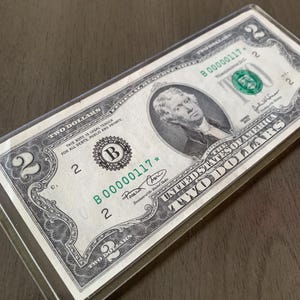 May include: A United States two-dollar bill, featuring a portrait of Thomas Jefferson. The bill is green and black, with the text "TWO DOLLARS" prominently displayed. The bill is in a clear protective sleeve.
