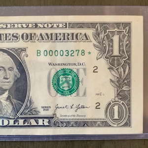 May include: A United States one-dollar bill, featuring a portrait of George Washington. The bill includes the text "RESERVE NOTE", "STATES OF AMERICA", "WASHINGTON, D.C.", and "DOLLAR". The bill is protected by a clear plastic sleeve.