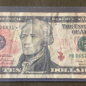 May include: A United States ten-dollar bill, featuring a portrait of Alexander Hamilton. The bill is encased in a clear plastic sleeve. The text "TEN DOLLARS" is visible, along with the seal of the United States.