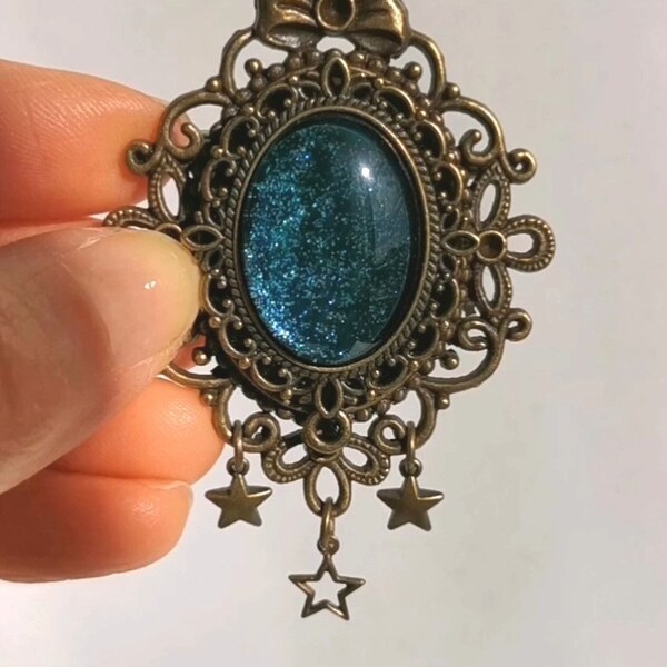 Broche ovale, en métal bronze avec breloques étoile, cabochon de verre, coloris bleu-vert, nuance "boréale", inspiration féérique, cosmique