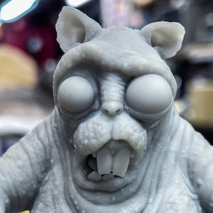 Könnte beinhalten: Eine graue, 3D-gedruckte Figur eines Wesens mit großen Augen, einem breiten Mund und einem zahnigen Grinsen. Das Wesen hat eine strukturierte, faltige Oberfläche und wirkt wie ein groteskes, karikiertes Tier.