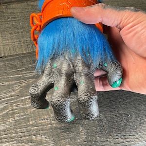 May include: A detailed, close-up view of a monster hand prop. The hand is gray with green spots and green painted nails. It has blue fur around the wrist and is adorned with an orange band and chain. An orange head is visible above the wrist.