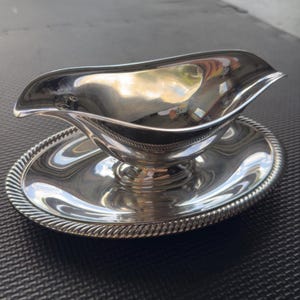 May include: A silver gravy boat with a matching saucer. The gravy boat has a curved spout and a handle. The saucer has a raised edge with a rope design. The set is reflective and appears to be made of metal.
