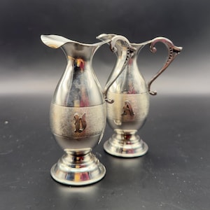 May include: Two silver-colored, small pitchers with ornate handles and bases. Each pitcher has a band with a letter on it. One pitcher has the letter "D" and the other has the letter "A". The pitchers have a shiny, reflective surface.