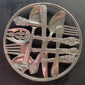 May include: A silver-colored, circular trivet featuring an arrangement of vintage silverware. The design includes forks, spoons, and knives, with ornate handles and a beaded border. The trivet is likely made of metal and is designed to protect surfaces from hot dishes.