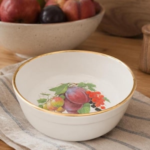 May include: Cream-colored ceramic bowl with a gold rim, featuring a fruit illustration of plums, red currants, and apples. The bowl is set on a striped cloth, with other bowls of fruit in the background.