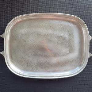 May include: Silver-toned oval serving tray with two handles. The tray has a slightly textured surface and a decorative floral pattern. The handles are rectangular with a curved design. The tray is made of metal.